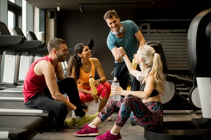 Vier energieke vrienden in sportkleding ontspannen en kletsen op de vloer van een sportschool, glimlachend na hun training. Op de achtergrond staan loopbanden opgesteld, die een levendige, motiverende sfeer uitstralen.